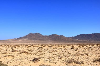 Arka planda yüksek volkanik dağlar olan çakıllı, tozlu bir yol. Jandia, Morro Jable, Fuerteventura, İspanya 'daki Kanarya Adaları