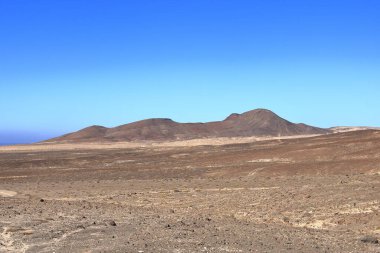 Arka planda yüksek volkanik dağlar olan çakıllı, tozlu bir yol. Jandia, Morro Jable, Fuerteventura, İspanya 'daki Kanarya Adaları