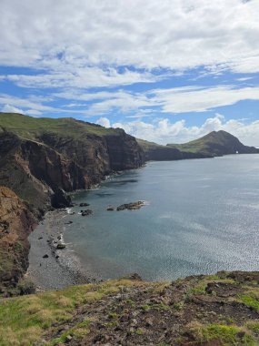 Ponta de So Loureno 'dan Madeiras dağlarına doğru bir manzara. Kırmızı volkanik toprak, sakin Atlantik suları ve bulutlu gökyüzünün altındaki uzak yeşil tepeler..