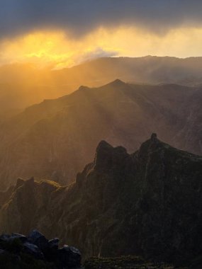 Altın güneş Pico do Arieiro 'nun üzerindeki bulut katmanlarını yararak dağın dramatik siluetini ortaya çıkarıyor ve dağlık araziyi sıcaklık ve ışıkla dolduruyor..