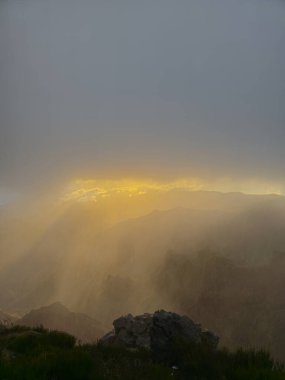 Güneş ışınları Pico do Arieiro 'nun üzerindeki bulutları delip geçer. Tepeler boyunca altın bir ışık saçar ve Madeira dağlarında sıcak, sakin bir atmosfer yaratır..