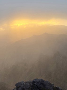 Güneş ışınları Pico do Arieiro 'nun üzerindeki bulutları delip geçer. Tepeler boyunca altın bir ışık saçar ve Madeira dağlarında sıcak, sakin bir atmosfer yaratır..