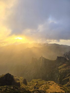 Pico do Arieiro 'nun üstündeki bulutların arasından süzülen altın güneş ışığı heybetli zirvelerin üzerine sıcak bir parıltı saçıyor ve sakin bir dağ kaçışı yaratıyor..