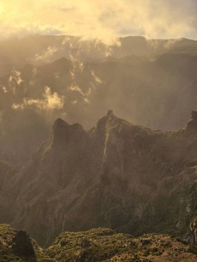 Pico do Arieiro 'nun üstündeki bulutların arasından süzülen altın güneş ışığı heybetli zirvelerin üzerine sıcak bir parıltı saçıyor ve sakin bir dağ kaçışı yaratıyor..