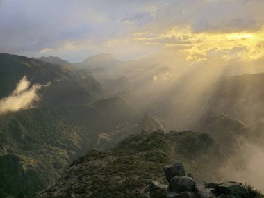 Pico do Arieiro 'nun engebeli tepelerinde güneş bulutları yarıp geçerken sıcak bir parıltı yayılıyor dramatik Madeira dağ manzarası üzerine sıcaklık saçıyor..