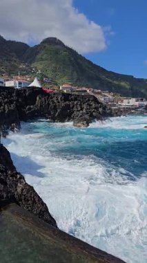 Porto Moniz, Madeira, Portekiz. Atlantik dalgaları, volkanik dağlar ve dramatik kıyı ışığı tarafından çerçevelenmiş bir sahil köyünün yanındaki siyah lav havuzlarına çarpar..