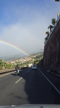 Portekiz 'in Madeira kentinin tepelerinde, sürücüler Funchal yakınlarındaki manzaralı bir otoyolda yürürken canlı bir gökkuşağı kemeri görülüyor. Şehir ve yollarla harmanlanmış nadir bir doğa manzarası..