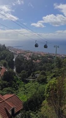 Kablolu araba Funchal, Madeira, Portekiz 'deki yemyeşil tepeler ve çatılar üzerinde süzülüyor. Atlantik kıyıları, tropikal ormanlar ve aşağıdaki şehrin panoramik manzaralı manzaralı bir yolculuk..