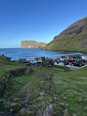 Faroe Adaları 'ndaki Tjrôk kıyı köyü dramatik dağ manzarasıyla çevrilidir ve açık deniz ve ikonik bazalt deniz yığınlarına tepeden bakar..
