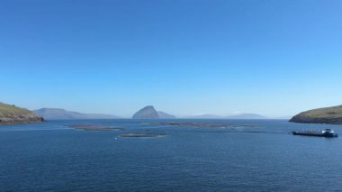Somon balığı kafesleri Faroe Adaları 'ndaki çimenli bazalt kayalıklarının altında sakin bir fiyortta yüzer ve bulutsuz yaz gökyüzü altında sürdürülebilir Kuzey-Atlantik su kültürünü resmeder..