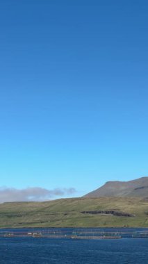 Uzak mavi sularda, Faroe Adaları 'nın kuzey Atlantik kıyılarında açık bir yaz gününde engebeli, otla kaplı uçurumların altında açık somon balığı çiftliği işleten servis botu..