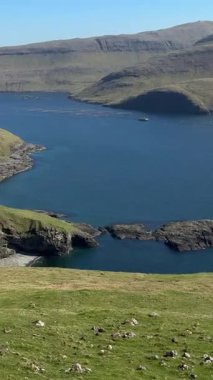Bur yakınlarındaki Rocky Sahili, Vgar Adası, Faroe Adaları. Engebeli kıyı şeridi ve açık kuzey gökyüzünün altındaki yeşil tepelerle çevrili derin mavi Atlantik suları..