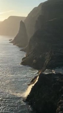 Günbatımı Faroe Adaları 'ndaki Vgar Adası' nın kayalıklarını aydınlatır ve Trllkonfingur denizin üzerinde yükselir. Efsanevi bir deniz yığını altın Atlantik ışığında parıldıyor..