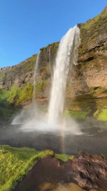 Seljalandsfoss Şelalesi İzlanda 'da görülebilir bir gökkuşağı ile sisin içinde şekilleniyor. Parlak mavi gökyüzü, gür kayalıklar ve çarpan su..