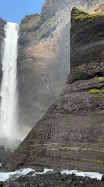 Hekla volkanı yakınlarındaki Hifoss şelalesi, İzlanda. Yosunlu ön planda dikey bir düşüş, karanlık volkanik kayalıklar, ve parlak bir yaz gökyüzünün altında kristal sprey.