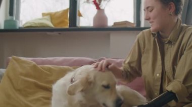 Protez kollu mutlu genç bir kadın evdeki kanepede otururken güzel golden retriever köpeğini okşuyor.