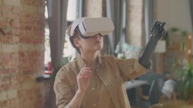 Beyaz bir kız, protez koluyla oturma odasında duruyor ve VR kulaklıkla video oyunu oynarken havaya dokunuyor.