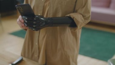 Protez kollu bir kızın evde mutfak masasının başında durup akıllı telefonuyla yemek fotoğrafı çekerken görüntüsü.