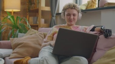 Kablosuz kulaklıklı, protez kollu bir kız evdeki kanepede oturuyor ve bilgisayardaki video görüşmesinde sohbet ediyor.