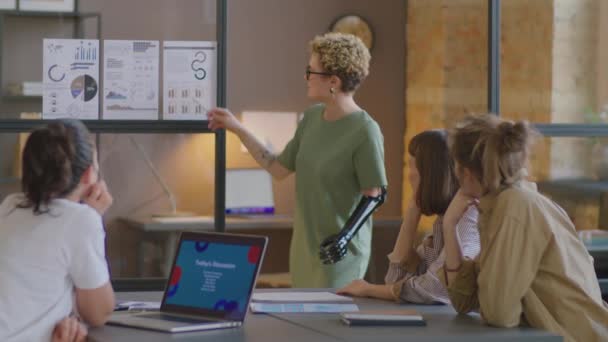 年轻女子 带着假臂 一边解释贴在玻璃幕墙上的纸片的商业图表 一边向同事们讲解 — 图库视频影像