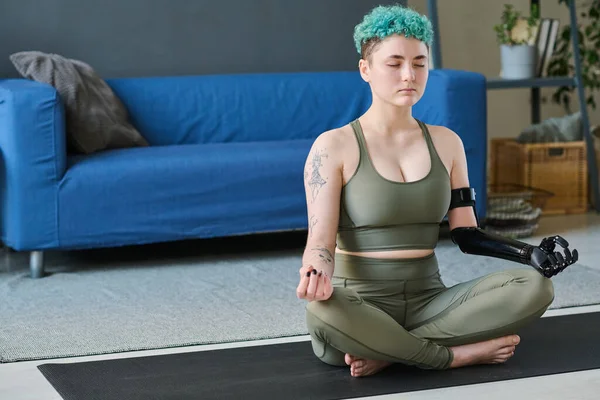 Young woman with prosthetic arm meditating in lotus position with her eyes closed in the room