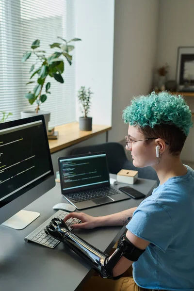Vertical image of young programmer with prosthetic arm working with computer codes on computer at her workplace at home