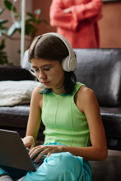 坐在房间里的一个戴无线耳机的少女 专心致志地在笔记本电脑上进行在线交流 — 图库照片