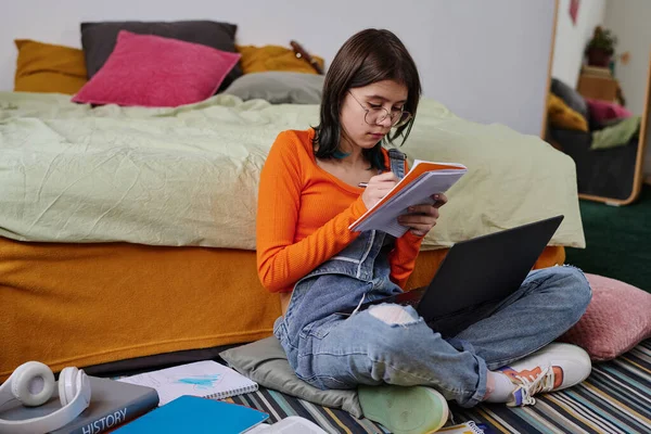 stock image Teenage girl sitting on the floor with laptop and making notes in notebook to prepare for her homework