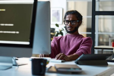 Gözlüklü genç müteahhit bilgisayardaki işine odaklanmış bilişim ofisinde masada oturuyor.