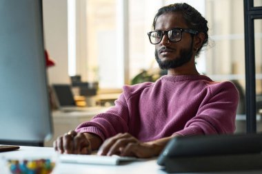 Gözlüklü ciddi bir genç programcı bilgisayarla bilgisayarında çalışıyor.