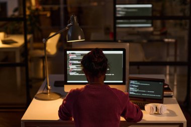 Son teslim tarihi boyunca bilişim ofisinde çalışan genç programcının arka görünüşü, monitör ve daktilo kodlarının önünde işyerinde oturuyor