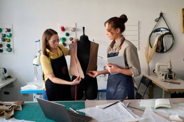 Genç terzi yardımcısı, her ikisi de mankenin önünde dikilirken bitmemiş deri yelek almasına yardım ediyor.