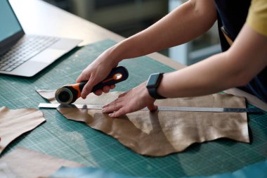 Genç bayan terzinin elleri, atölyede ya da stüdyoda yeni kıyafet malzemesi yaratmak için çalışırken masada bej bir deri parçası kesiyor.
