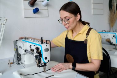 İş yerinde atölye veya atölyede elektrikli dikiş makinesi ile otururken profesyonel kadın tabakçı yeni deri kıyafetler üretiyor.