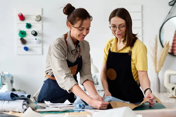 テーブルに立って作業服の2つのプロの女性のタンナーとクライアントのための新しいアパレルを縫製するための革の一部を選択 — ストック写真