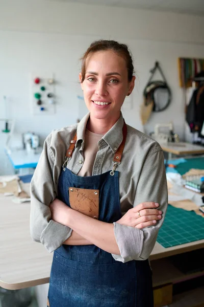 Young successful female leather worker and owner of atelier in apron looking at camera with smile while standing by her workplace