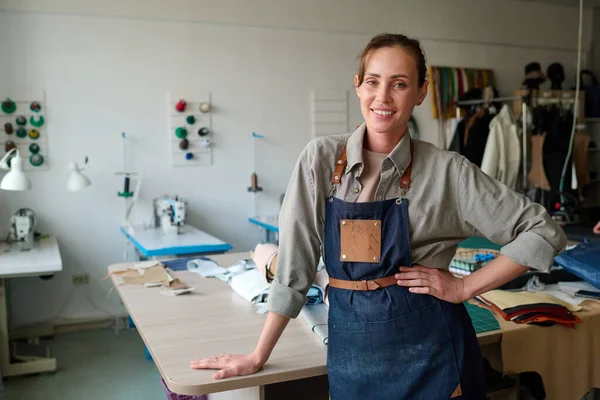 stock image Happy young craftswoman in apron standing by workplace in atelier with large table and group of desks with electric sewing machines