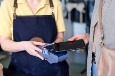 Genç bayan müşterinin kart okuyucuya karşı akıllı telefonu tutarken dükkan asistanının önünde durup satın almak için para ödemesi.