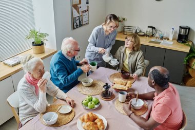 Genç bakıcıların, yaşlı insanlar masada otururken ve kahvaltı yaparken, onlara süt içirmelerinin yüksek açılı görüntüsü.