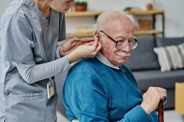 老人ホームで高齢者の介護をしながら補聴器を身につけるのを手伝っている若い介護者 — ストック写真
