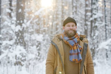 Sıcak giyimli genç sakallı adamın portresi. Kış ormanında kameraya gülümsüyor.