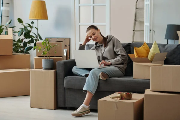 若い女性がソファの上に座って梱包箱の間で 新しいアパートを借りるために探しています — ストック写真