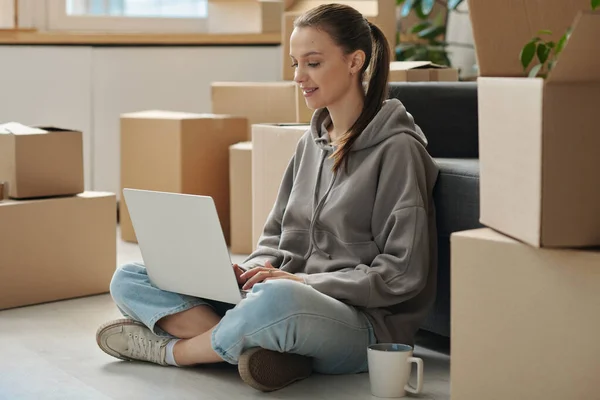 stock image Young woman using laptop to rent a new apartment while sitting on the floor among packed boxes during relocation