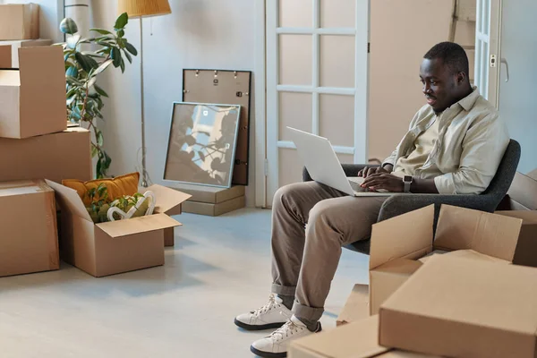 アフリカ系アメリカ人の若い男がアームチェアに座ってノートパソコンでオンラインで作業し 移転中に箱の中の部屋に座って — ストック写真