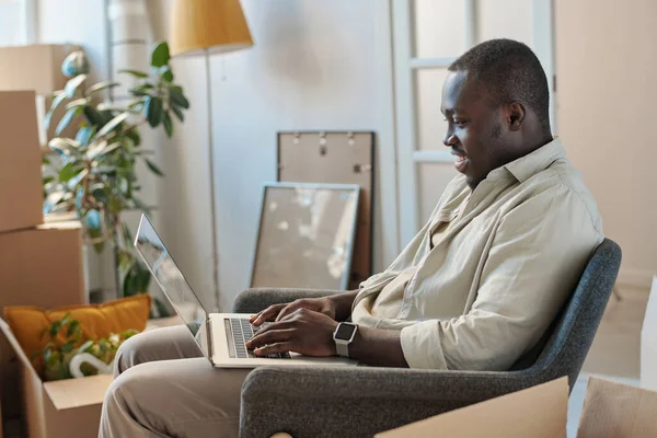 アフリカ系アメリカ人の男性は 部屋のアームチェアに座っている間 ラップトップ上でオンライン通信を持っています — ストック写真