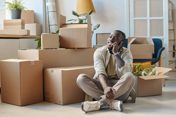 アフリカ系アメリカ人の男性は 段ボール箱の間の床に座って 新しい家に移動中に携帯電話で話しています — ストック写真