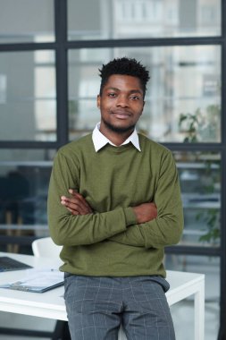 Genç Afro-Amerikan iş adamının portresi. Kollarını iş yerinde kavuşturup kameraya bakıyor.