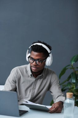 Kulaklıklı Afro-Amerikan bir adamın bilgisayarda oturduğu ve internette çalıştığı dikey görüntü.