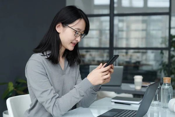 stock image Asian businesswoman communicating online on her smartphone while working at her workplace with laptop