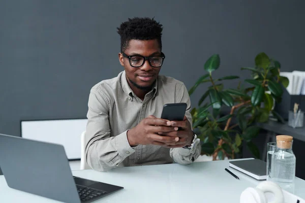 アフリカ系アメリカ人のビジネスマンがノートパソコンを持って職場に座っているスマートフォンでオンラインで働いています — ストック写真